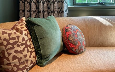 A tan sofa styled with three decorative pillows, including a geometric brown and beige pillow, a deep green velvet pillow, and a round red pillow with intricate floral embroidery. The sofa is set in front of a window framed by elegant black and white damask-patterned curtains. The natural light highlights the soft texture of the upholstery and vibrant colors of the decor.