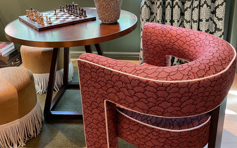 A cozy corner featuring a circular wooden table set with a chessboard and a textured decorative vase. Surrounding the table are a plush red chair with a scalloped pattern and white piping, and fringed beige ottomans. The scene is completed with green walls, patterned curtains, and a comfortable, inviting atmosphere.