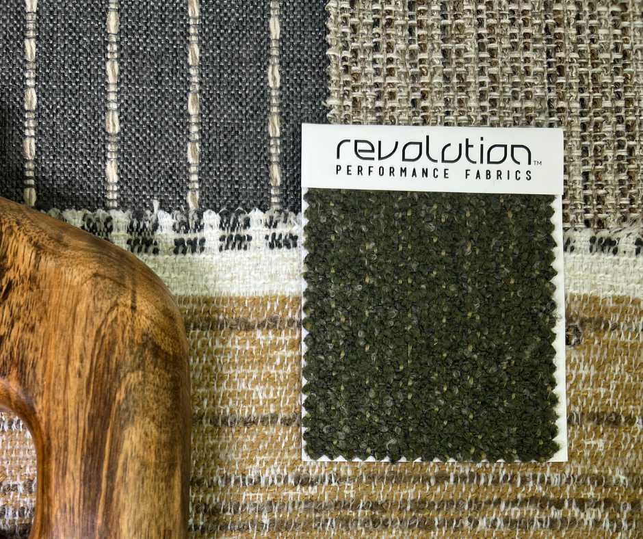 Close-up of a Revolution Performance Fabrics swatch card featuring a dark green boucle texture, placed on a layered backdrop of woven outdoor textiles in earthy tones with a carved wooden accent.