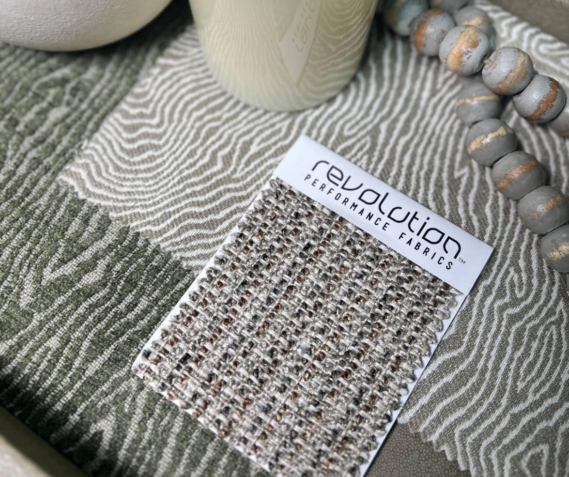 Revolution Performance Fabrics swatch card in a neutral, chunky weave pattern styled on top of a soft green zebra-like patterned fabric with ceramic accessories and wooden beads.