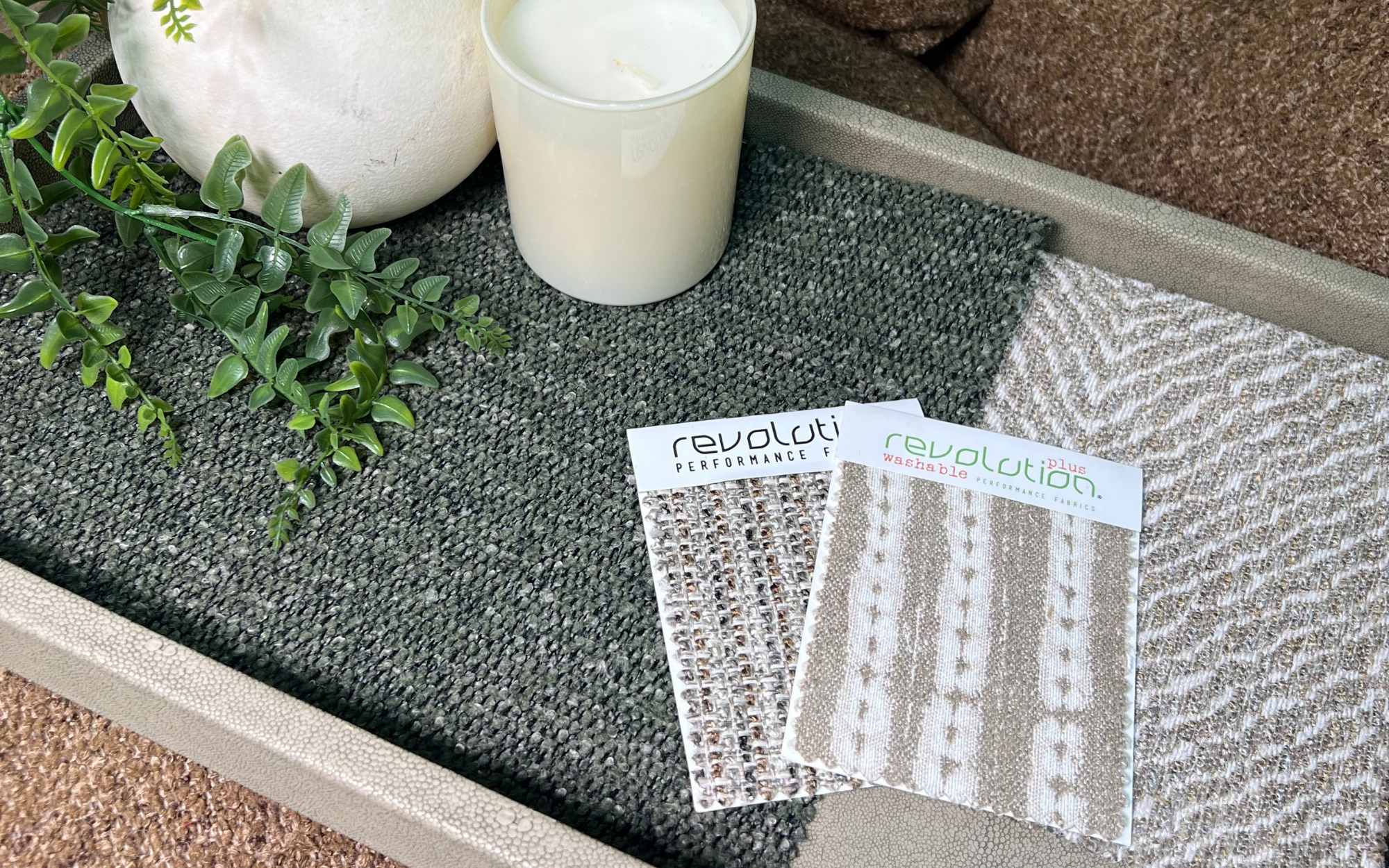 A styled tray display with two Revolution fabric swatches—one chunky neutral weave and one striped boucle—laid over a textured green fabric, with a plant, candle, and ceramic accessories adding natural accents.