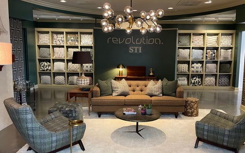 A stylish showroom featuring a mid-century modern aesthetic with a tufted tan sofa decorated with green textured throw pillows. The backdrop showcases a wall of cubby shelves filled with neatly folded patterned fabrics in various designs and neutral tones. The room includes plaid upholstered chairs, a round coffee table with magazines and plants, a table lamp with a patterned base, and a modern chandelier with multiple globe lights, creating a cozy and elegant ambiance.