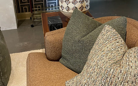 A close-up of a tan upholstered sofa with green and multi-colored textured throw pillows adding depth and comfort to the design. In the background, a mid-century style wooden side table holds a mosaic-patterned lamp and decor items. The setting is completed by a neutral rug and geometric wall patterns, creating a sophisticated and cozy ambiance.