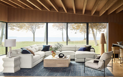 A modern and spacious living room with a cozy sectional sofa in white, adorned with accent pillows in navy and brown. A light wood coffee table holds decorative objects, and the room features a contemporary armchair and textured table lamp. Large floor-to-ceiling windows frame a scenic outdoor view of trees and a body of water, complemented by exposed wood beams and a warm, minimalist design.
