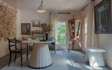 A bright and eclectic room with a modern, round wooden dining table surrounded by a mix of vintage and contemporary chairs. The space is adorned with unique artwork, including a textured wall hanging, a portrait, and a monochromatic landscape painting. A wooden display cabinet with angled legs holds decorative items, while natural light streams through a large window, highlighting a cozy boucle stool and an arrangement of dried flowers on the table.