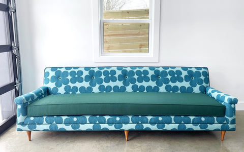 A vibrant mid-century modern sofa with a bold blue floral-patterned upholstery and a solid teal seat cushion. The sofa features clean lines, tapered wooden legs, and button tufting on the backrest, set against a bright white wall with a window. The minimalistic setting emphasizes the sofa's striking design and retro-inspired aesthetic.