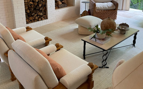 **ALT text:** A cozy and inviting living room with white upholstered armchairs featuring wooden frames and soft terracotta accent pillows. The space includes a woven accent chair, a modern coffee table with a marble top and black wrought-iron legs, styled with a potted plant and decorative items. A white brick fireplace with stacked firewood and neutral-toned decor enhances the warm, relaxed atmosphere.