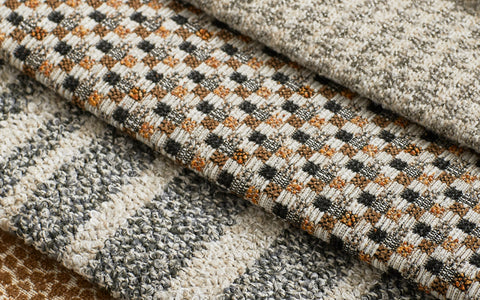 A close-up of stacked upholstery fabric swatches showcasing various textures and patterns. The designs include a small-scale checkered pattern in warm brown and black tones, a speckled weave with neutral shades, and a boucle-style fabric with gray and cream tones. The intricate details and rich textures highlight the versatility and craftsmanship of the fabrics.