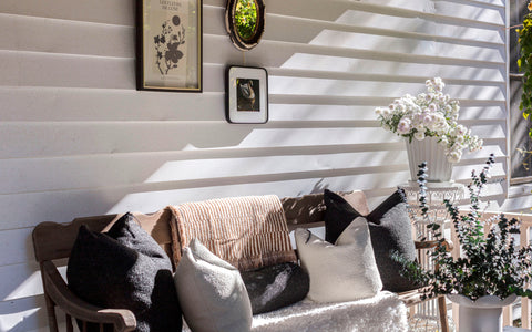 A charming outdoor bench set against white horizontal siding, decorated with cozy throw pillows in shades of black, white, and cream, and a striped beige blanket. The bench is surrounded by decorative touches, including a wall adorned with framed artwork and a mirror, and a white pedestal holding a pot of delicate white flowers. Soft natural light casts shadows, creating a serene and inviting setting.