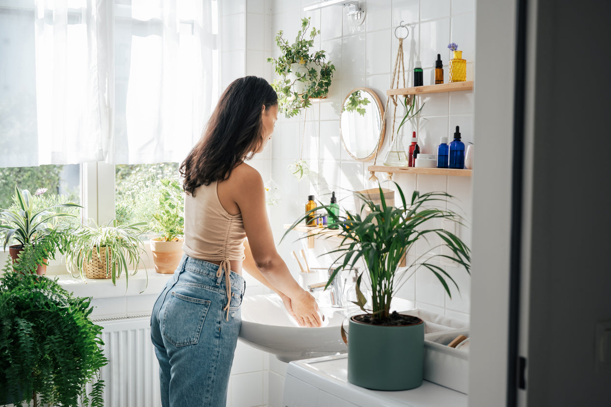 A woman in a sunlit bathroom surrounded by vibrant indoor plants, washing her hands at a white sink under a mirror, evoking a clean, biophilic environment.