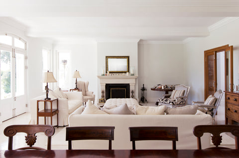 A bright and elegant living room featuring cream-colored sofas and armchairs arranged around a tufted ottoman. A classic fireplace with a gilded mirror above serves as the focal point, flanked by traditional side tables with decorative lamps. The space is adorned with wooden furniture, striped accent chairs, and natural light streaming in through large windows and French doors, creating a refined and inviting atmosphere.
