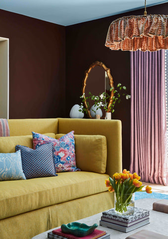 A vibrant living room with a mustard-yellow sofa adorned with colorful patterned throw pillows in pink, blue, and geometric designs. A marble coffee table holds a vase of bright orange tulips and stacked books. The room features rich brown walls, a gold-framed mirror with greenery, and mauve curtains, all complemented by a statement fabric pendant light above.