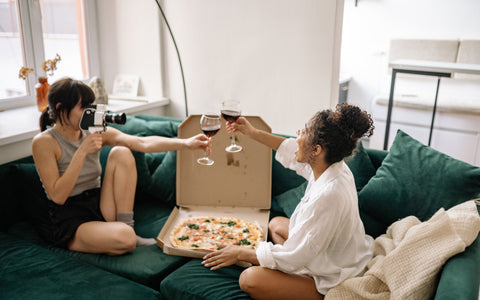 Two women cheering their glasses of wine enjoying pizza on a green couch
