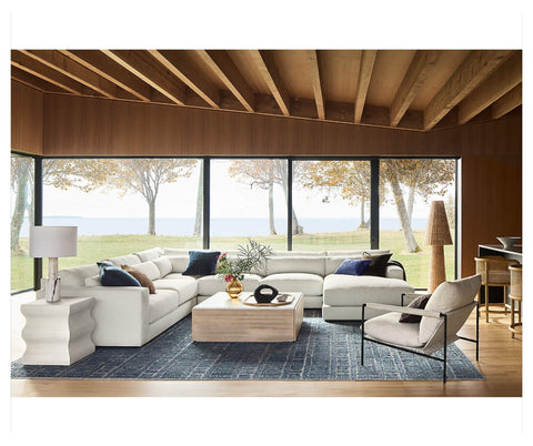 A modern living room with large windows overlooking a scenic outdoor view, featuring a white sectional sofa upholstered in Revolution Performance Fabrics, a coffee table, and a cozy armchair.