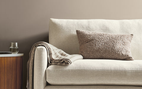 A sleek and modern beige sofa with clean lines, featuring a textured taupe throw pillow and a striped throw blanket draped over the armrest. The sofa is paired with a mid-century style wooden side table holding a glass and small decorative items. The neutral-toned backdrop creates a calming and minimalist aesthetic.