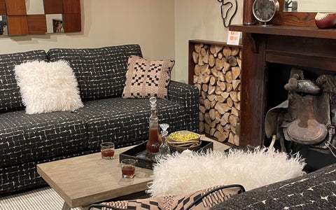 A cozy living room with a dark patterned sofa featuring a white geometric design. The sofa is adorned with a fluffy white pillow and a beige patterned throw pillow. The space includes a wooden coffee table holding a decanter, glasses, and a basket of snacks, complemented by a rustic decor with a fireplace stacked with logs and a vintage saddle on display.