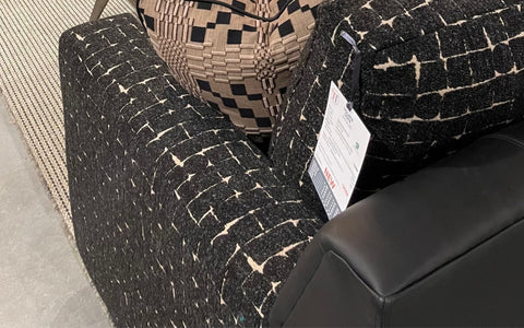 A close-up of a dark patterned sofa with a white geometric design, showcasing the detailed texture and a beige patterned throw pillow. A price tag hangs from the sofa armrest, with the corner of a black leather armrest visible in the frame. The scene is set on a neutral-toned woven rug.