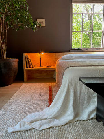 A serene and minimalist bedroom with a low wooden platform bed dressed in white linens that cascade onto a textured cream rug. A built-in wooden nightstand glows softly with a small lamp, holding books and a dish. The room features dark walls, a large potted tree for a natural touch, and a window with a view of lush green trees, creating a calm and inviting atmosphere.