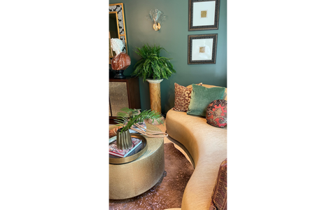 A sophisticated living space featuring a curved beige sofa adorned with patterned and solid throw pillows, including a green velvet and a red embroidered design. A round, gold-toned coffee table holds a decorative vase with fern leaves and magazines, complementing the elegant decor. The backdrop includes a tall fern on a pedestal, framed artwork on a green wall, and a sculpture atop a cabinet, creating a luxurious and curated ambiance.